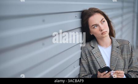 Erschöpfte professionelle Frau, die an der Wand ruht und das Smartphone mit müdem Ausdruck packt Stockfoto