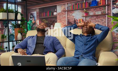 afroamerikanisches Paar, das sich auf der Couch entspannt, sich extrem müde fühlt und unter Druck arbeitet. Schläfrige Menschen, die mit Burnout und Arbeitsbelastung zu kämpfen haben, Freiberufler mit Angst. Kamera B.. Stockfoto