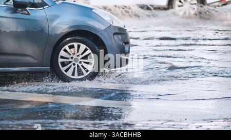 Graues Auto, das nach starkem Regen auf einer überfluteten Straße navigiert und Wasserspritzer aufwirft, während es durch Pfützen auf nassem Asphalt fährt Stockfoto