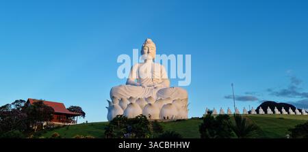 Während das letzte Licht des Tages der Dämmerung weicht, sitzt eine ruhige und majestätische weiße Buddha-Statue friedlich auf einem grünen Hügel. Stockfoto
