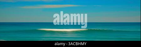 Eine ruhige Szene, in der eine sanfte Welle am Sandstrand bricht, während die Sonne untergeht und ein warmes Licht auf Wasser und Sand wirft. Das Wasser ist ein lebhaftes Türkis Stockfoto