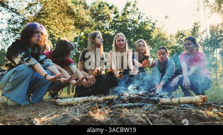 Junge Studenten, die sich am knisternden Lagerfeuer tummeln, Hände erwärmen und Kameradschaft beim Camping-Abenteuer im Wald teilen Stockfoto