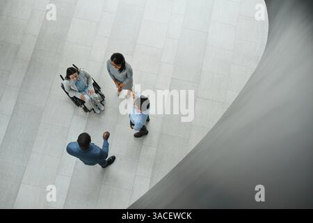 Grafische Ansicht von oben nach unten auf junge Geschäftsfrau im Rollstuhl, die mit verschiedenen Gruppen von Personen in der Lobby des Büros und im Kopierbereich spricht Stockfoto
