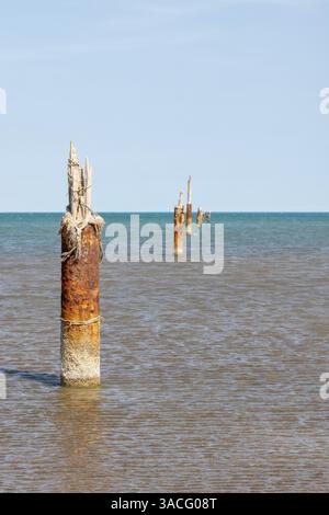 Verwitterte rostige Pfosten erheben sich aus dem flachen Meer und verkörpern Geschichte inmitten der ruhigen Küstenlandschaft und des blauen Himmels. Vertikales Foto am Kaspischen Meer Stockfoto