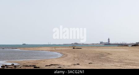 Ruhige Küstenszene mit ausgedehntem Sandstrand, ruhigem Wasser, einem entfernten Pier und einem Leuchtturm unter klarem Himmel. Küste des Kaspischen Meeres, Buzowna, Stockfoto