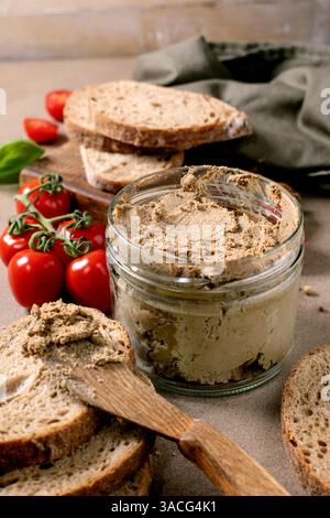 Hausgemachte Fleischpastete im Glas, serviert mit frischen Vollkornbrotscheiben, Kirschtomaten und Basilikumblättern. Rustikaler Holzlöffel und Schneidebrett. Stockfoto