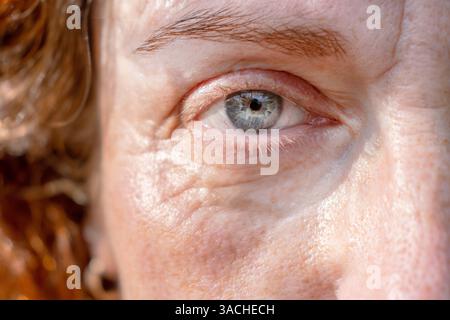 Detaillierte Nahaufnahme des rechten blaugrauen Auges einer ernsten rothaarigen Frau mittleren Alters, die unauffällig vor der Kamera liegt. Die Hälfte des Face Macro Shots. Fin Stockfoto