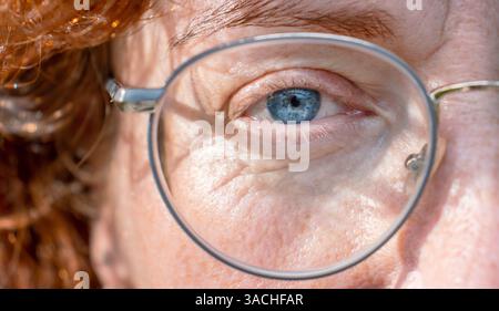 Detaillierte Nahaufnahme des rechten blaugrauen Auges einer ernsten rothaarigen Frau mittleren Alters mit einer Brille in Metallrahmen. Die Hälfte des Face Macro Shots. Fi Stockfoto