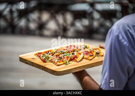 Nahaufnahme des Kellners, der Pizza zum Kunden im Restaurant im Freien brachte. Gourmet-Pizzascheiben auf Holzbrett als Teller unter Sonnenschein. Urlaubsresort Stockfoto