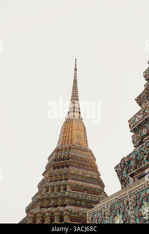 Im Wat Pho erheben sich goldene Türme in einen hellen Himmel, gekrönt von zarten Keramikblüten Stockfoto