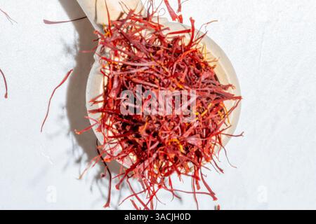 Rote Safranfäden im Holzlöffel, die auf weißem Marmorhintergrund verschüttet werden, hell und luftig mit Sonnenlicht und Schatten, Ansicht von oben nach unten Stockfoto