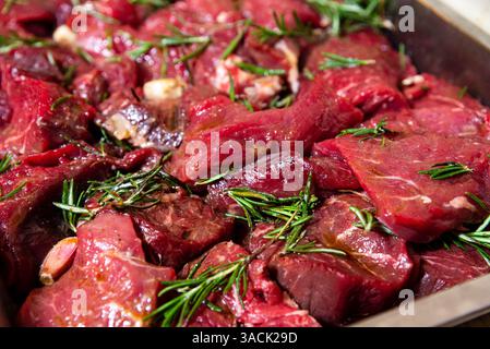 Nahaufnahme frischer roher Rindfleischstücke in einem Tablett, mariniert mit Olivenöl, Knoblauch und Rosmarin zum Kochen oder Grillen. Stockfoto