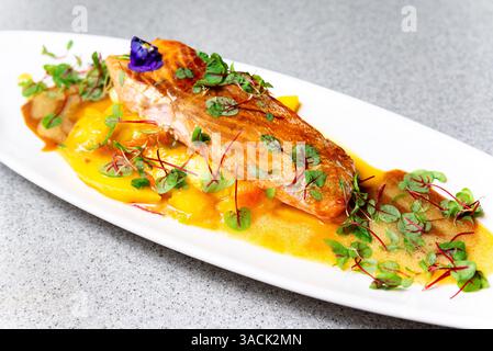 Überzogenes Gourmet-Lachsfilet, serviert auf lebendiger Mangohauce, garniert mit frischem Mikrogrün und einer essbaren Blume. Stockfoto