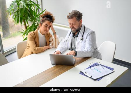 Hochwinkelansicht eines männlichen Arztes mit Testergebnissen bei belasteter Patientin über Laptop in der medizinischen Klinik Stockfoto