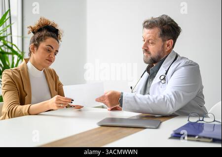 Männlicher Arzt zeigt und bespricht medizinische Testergebnisse mit trauriger Patientin am Schreibtisch in der medizinischen Klinik Stockfoto
