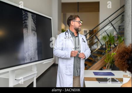 Männlicher Arzt im Laborkittel, der Wirbelsäulenröntgen über Computerbildschirm während der Arbeit im modernen Krankenhaus untersucht Stockfoto