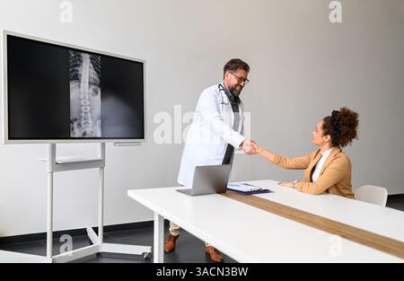 Männlicher Arzt mit Röntgenbild auf dem Computerbildschirm, der mit sitzender Patientin im Krankenhaus die Hände schüttelt Stockfoto