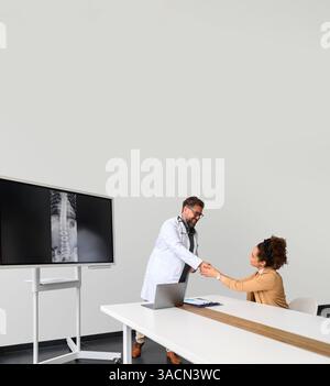 Patientin schüttelt die Hände und drückt Dankbarkeit gegenüber dem männlichen Arzt aus, während sie am Schreibtisch im Krankenhaus sitzt Stockfoto
