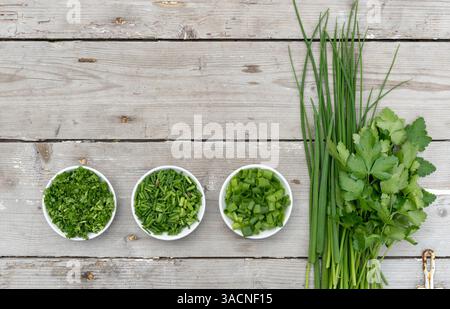 Frische Petersilie, Schnittlauch und Frühlingszwiebeln auf hellem hölzernem Hintergrund bündeln und schneiden, Stockfoto