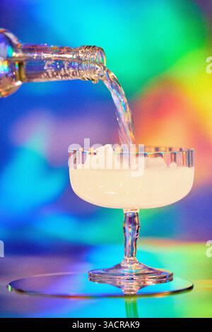 Trinken Sie das Gießen in Cocktailglas aus der Flasche auf farbenfrohen hellen Neonlichthintergrund, Filmkörnung hinzugefügt, selektiver Fokus Stockfoto