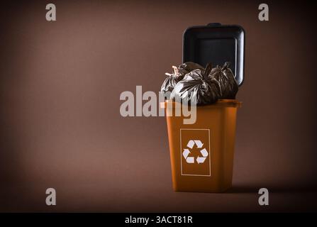 Recyclingkonzept für organische Abfälle mit einem braunen Abfallbehälter mit dem Recycling-Symbol gefüllt mit dunkelbraunen Beuteln auf braunem Hintergrund. Vorderansicht. Stockfoto