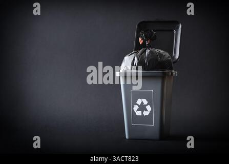 Allgemeines Abfallrecyclingkonzept mit einem grauen Recyclingbehälter mit einem Recyclingsymbol, gefüllt mit einem schwarzen Beutel auf grauem Hintergrund. Vorderansicht. Stockfoto
