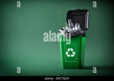 Recyclingkonzept mit grünem Abfallbehälter, gefüllt mit Glasbehältern mit Recycling-Symbol auf grünem Hintergrund. Vorderansicht. Stockfoto