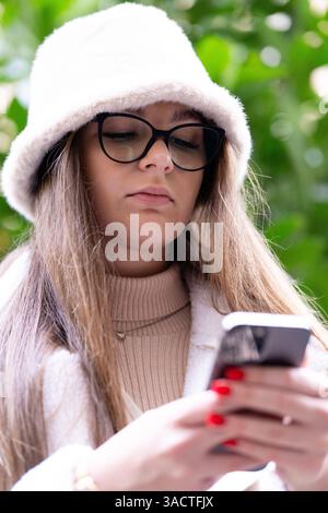 In einer lebhaften urbanen Umgebung konzentriert sich eine junge Frau in modernem Outfit auf ihr Smartphone, umgeben von üppigem Grün und dem Charme der Stadt, cre Stockfoto