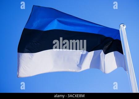Wunderschöne Flagge Estlands, die im Wind auf dem Fahnenmast gegen blauen Himmel mit Wolken winkt. Nahaufnahme der estnischen Nationalflagge, die an windigem Tag flattert. Stockfoto