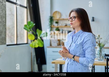 Porträt einer ernsten Frau mittleren Alters, die zu Hause aus dem Fenster blickt Stockfoto