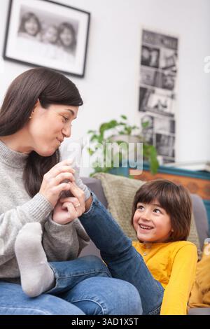Ein verspielter und liebevoller Moment, in dem die Mutter die Socken ihres Sohnes auszieht, während sie fröhliches Lächeln austauschen. Fängt emotionale Wärme und Verbundenheit ein. Stockfoto