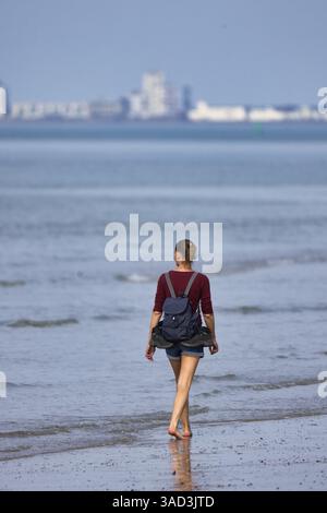 Hübsche junge Frau mit Rucksack und Shorts, die barfuß in der Sonne durch das flache Meer am Strand mit der Silhouette von Vlissingen geht Stockfoto