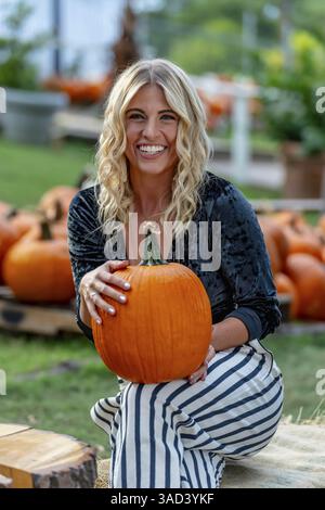 Ein atemberaubendes blondes Model posiert in einem lebhaften Kürbisfeld und trägt ein stilvolles Herbstoutfit mit einem Hauch von Halloween-Charme. Die Einstellung leuchtet warm Stockfoto