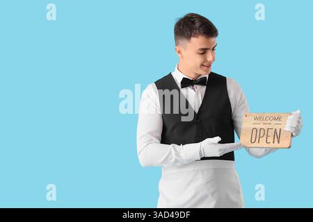 Junger männlicher Kellner, der zeigt, dass WIR EIN OFFENES Schild auf blauem Hintergrund haben Stockfoto