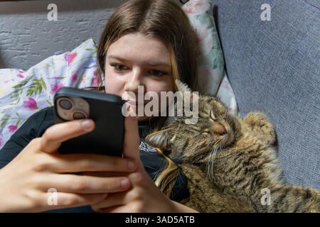 Ein Mädchen mit einer Katze, das zu Hause ein Selfie auf einem Smartphone macht. Die Katze liegt auf der Couch. Stockfoto