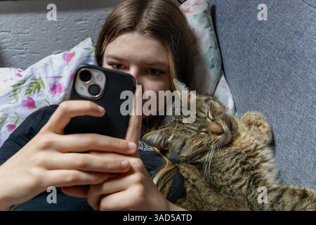 Ein Mädchen mit einer Katze, das zu Hause ein Selfie auf einem Smartphone macht. Die Katze liegt auf der Couch. Stockfoto