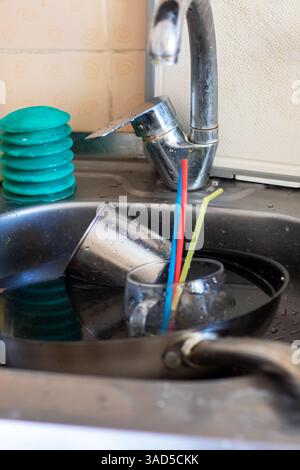 Eine Küchenspüle, die bis zum Rand gefüllt ist mit einem überwältigenden Stapel schmutzigem Geschirr, Utensilien und bunten Strohhalmen Stockfoto