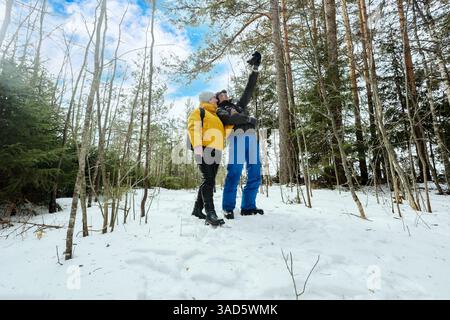 Ein reifes Paar steht nahe im schneebedeckten Wald, während der Mann die Kamera hochhebt, um Selfie zu machen, und die Frau blickt auf sie auf und genießt unbeschwerte Momente, während sie unterwegs ist Stockfoto