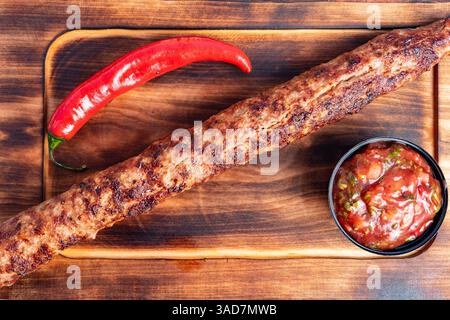 Saftiger Kebab liegt auf einem Holzbrett, begleitet von einer Schüssel mit würziger Salsa und einer lebendigen roten Chili-Pfeffer. Perfekt für ein schmackhaftes Essen zu jeder Zeit Stockfoto