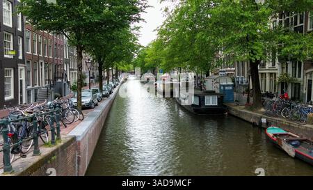 Eine malerische Kanalszene in Amsterdam, gesäumt von Bäumen und traditionellen niederländischen Gebäuden. Die Boote liegen am Wasser vor, und die Fahrräder stehen auf dem Parkplatz Stockfoto