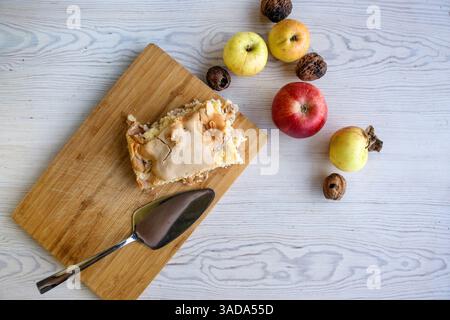 Ein Stück traditioneller Thanksgiving-Apfelzimtkuchen auf dem hölzernen Schneidebrett mit Äpfeln und Walnüssen auf dem weißen Tisch. Hausgemachte gebackene Obsttorte Stockfoto