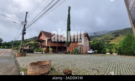 Eine malerische Landschaft mit einem charmanten zweistöckigen Backsteinhaus mit Holzbalkonen und einem gekachelten Dach, das sich vor einem üppig grünen Berg befindet. Stockfoto
