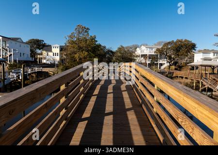 Eine sonnendurchflutete Holzstege führt durch ein ruhiges Viertel von Oak Island mit modernen Häusern an der Küste und einem klaren blauen Himmel. Stockfoto