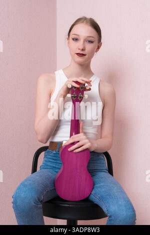 Porträt einer ruhigen jungen Musikerin mit aufrecht gehaltener Ukulele, die auf Barhocker sitzt und selbstbewusst in die Kamera blickt. Schöne Frau in lässiger cl Stockfoto