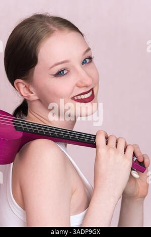 Porträt einer jungen Musikerin hält Ukulele am Hals und wirft Musikinstrument über die Schulter. Die schöne 18-jährige Frau schaut in die Kamera Stockfoto