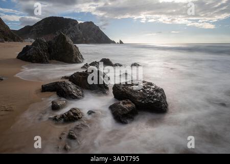 Lange Exposition von Wellen, die über Felsen am Adraga-Strand bei Sonnenuntergang mit Klippen im Hintergrund, sintra, portugal Stockfoto