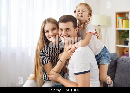 Glückliche Familie, die auf dem Sofa sitzt, lächelnde Eltern umarmt, während ihre Tochter sie von hinten umschließt. Stockfoto