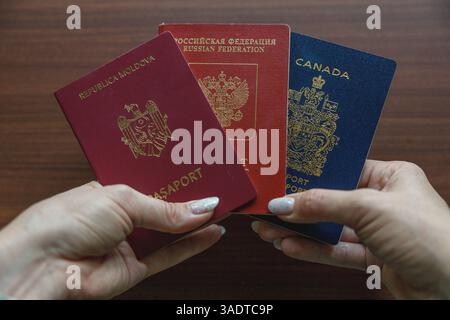 Vancouver, Kanada - 18. März 2025: Hände mit drei Pässen, die auf hölzerner Oberfläche gefächert sind – burgunderroter moldauischer, roter russischer und blauer kanadischer Pass Stockfoto