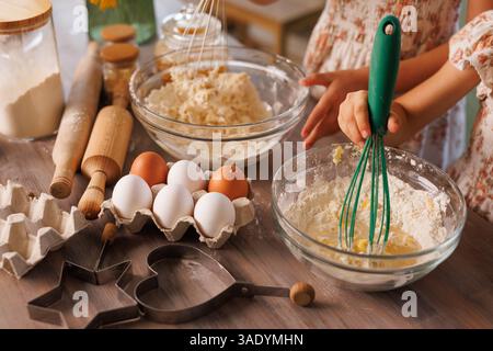 Kinder rühren Mehl und Eier in Glasschüsseln, während sie gemeinsam Kekse in einer warmen, rustikalen Küche backen. Stockfoto