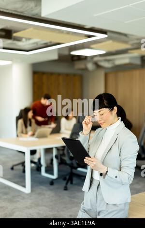 In einem modernen Büro verwendet eine fokussierte japanische Geschäftsfrau in einem stilvollen Anzug ihr Tablet, während die Teammitglieder im Hintergrund zusammenarbeiten und so eine Stockfoto